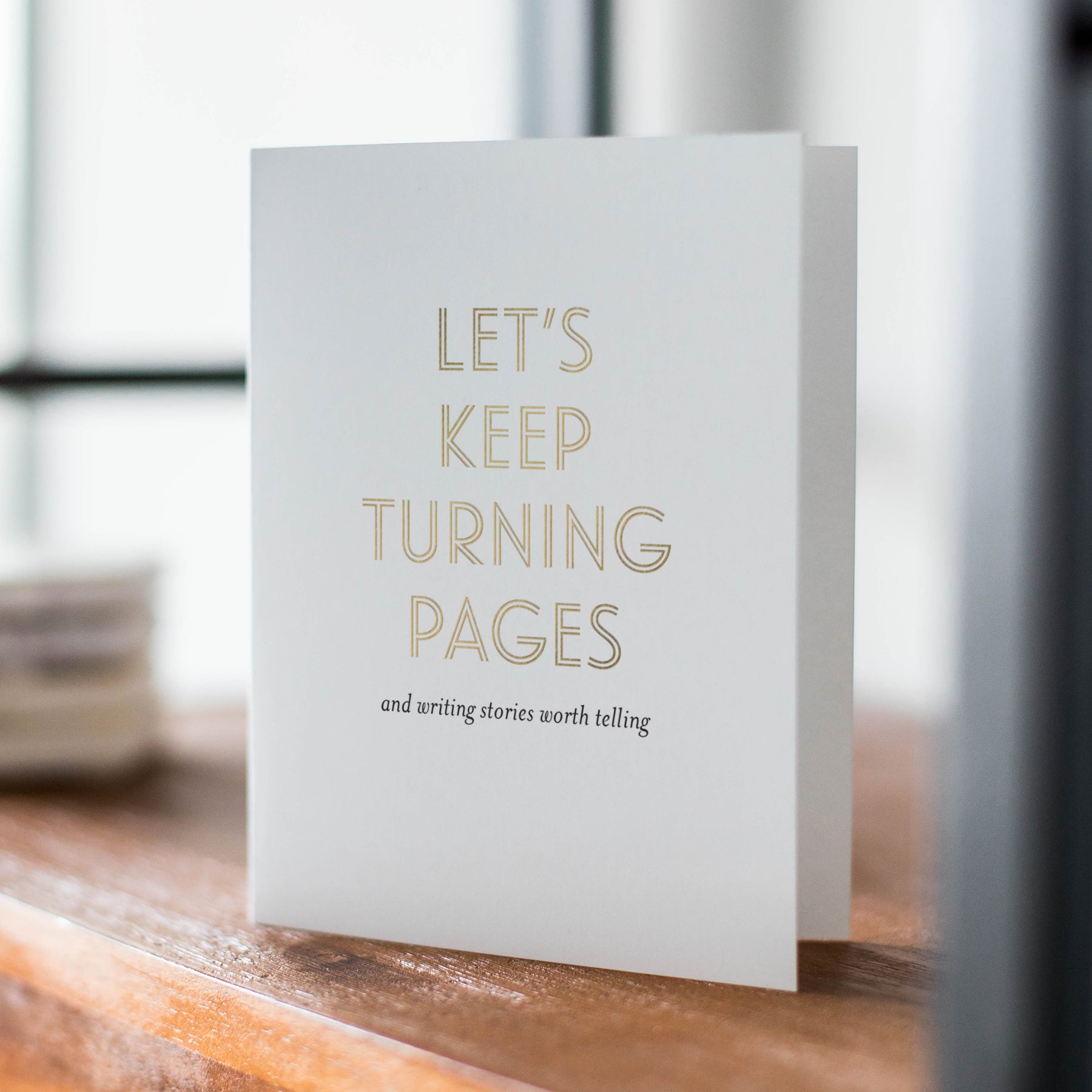 Greeting card with 'Let's keep turning pages and writing stories worth telling' text on a wooden surface.