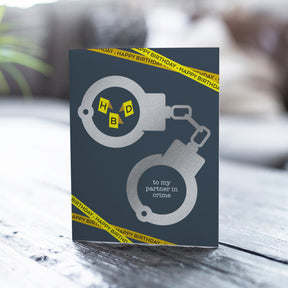 Greeting card with handcuffs design and 'to my partner in crime' text on a wooden surface.