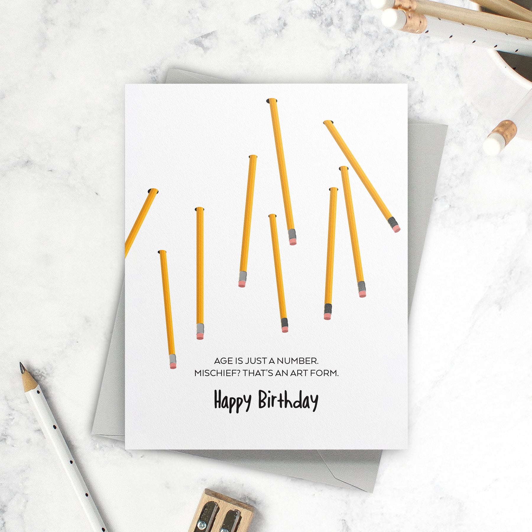 Front view of birthday card with yellow pencils on a desk surface. Card reads "Age is just a number. Mischief? That's an art form." 