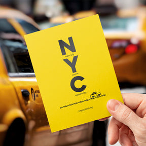 Yellow card with 'Naturally NYC' text held in front of a yellow taxi.