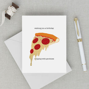 Greeting card with pizza slice illustration and text on a gray surface with an envelope and pen.
