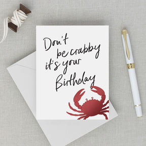 Birthday card with red crab illustration and text 'Don't be crabby it's your birthday' on a gray surface with an envelope and pen.