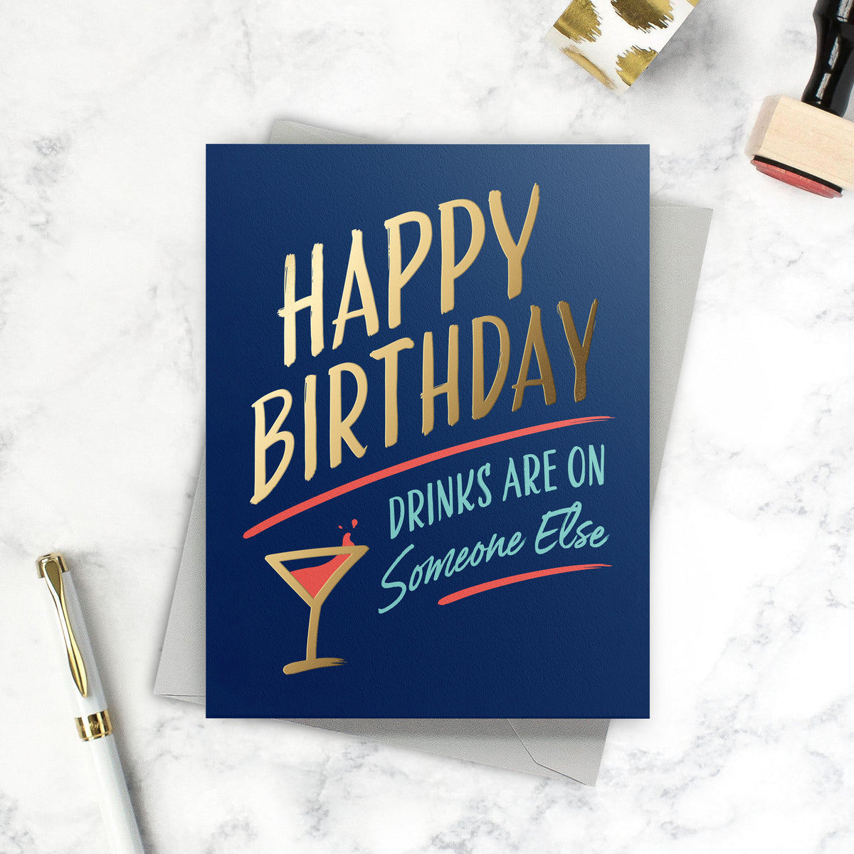 Lifestyle photo featuring a birthday card with navy blue background that says "Happy Birthday – Drinks are on someone else". 