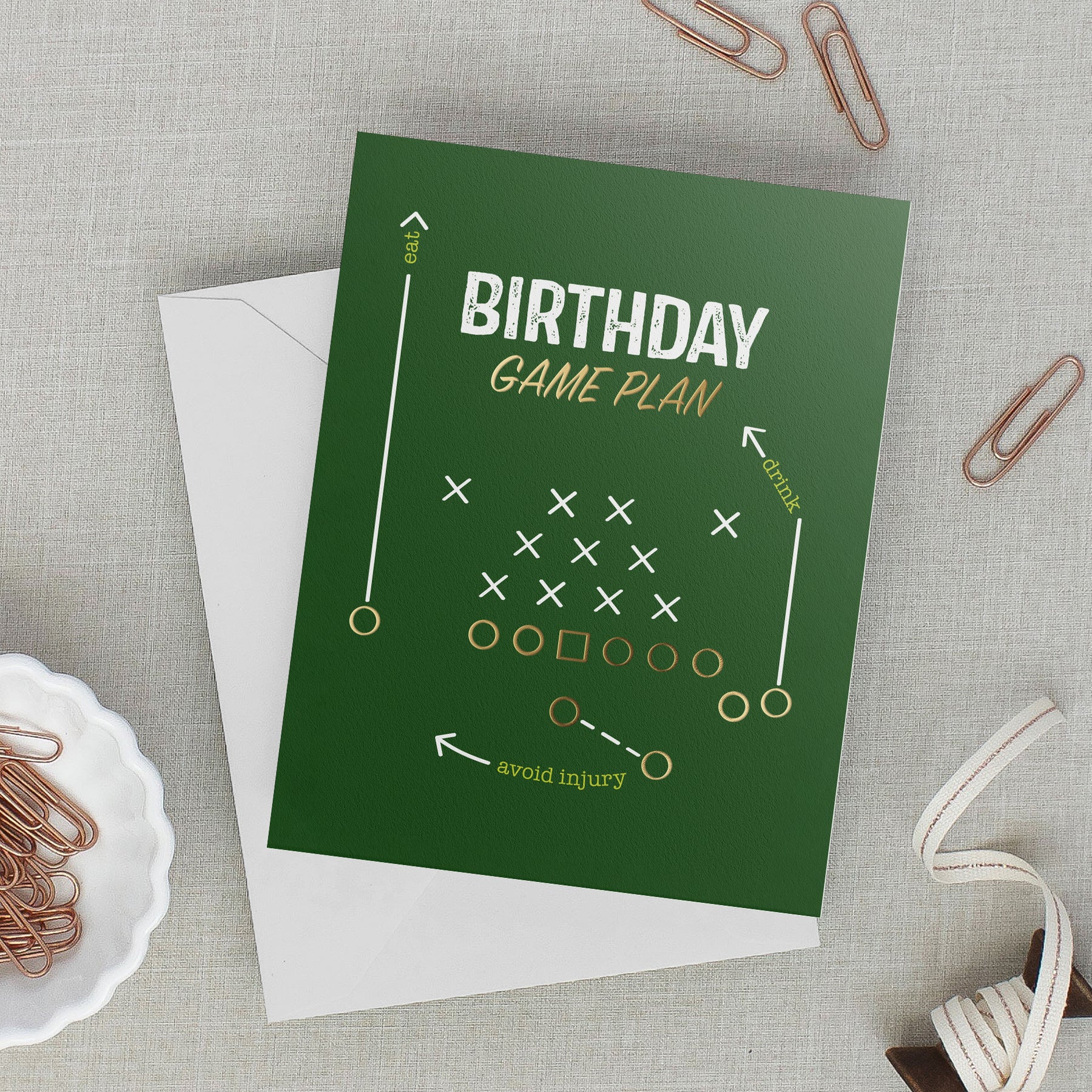 Green football birthday card reading “Birthday Game Plan: eat, drink, avoid injury,” with gold and white playbook symbols, resting on a light gray envelope on gray linen.