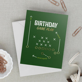 Green football birthday card reading “Birthday Game Plan: eat, drink, avoid injury,” with gold and white playbook symbols, resting on a light gray envelope on gray linen.