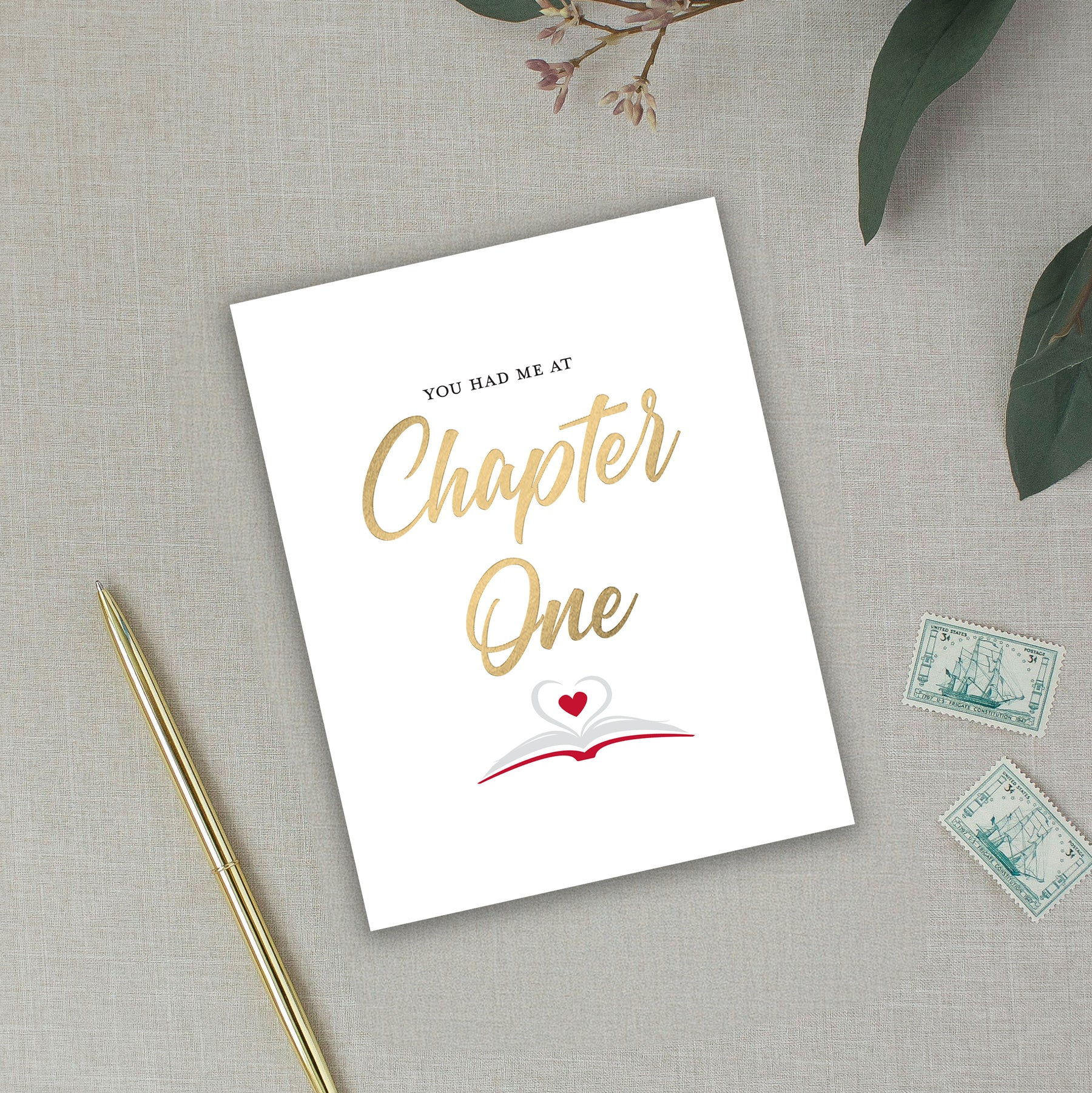 Greeting card with 'You had me at Chapter One' text on a textured surface with a pen and stamps.