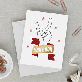 Greeting card with hand gesture and 'You Rock' text on a gray surface with paper clips.