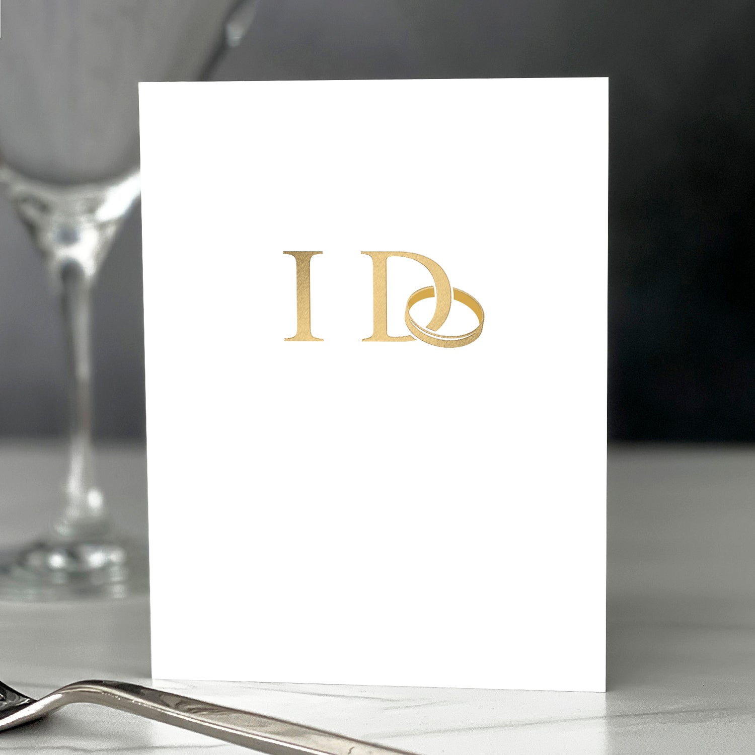 White card with gold monogram on a table setting with a glass and fork.